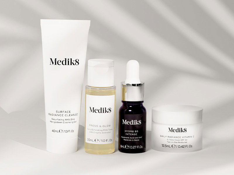 Four travel-sized skincare products lined up against a light gray background with soft shadow overlays. From left to right: a white squeeze tube of 