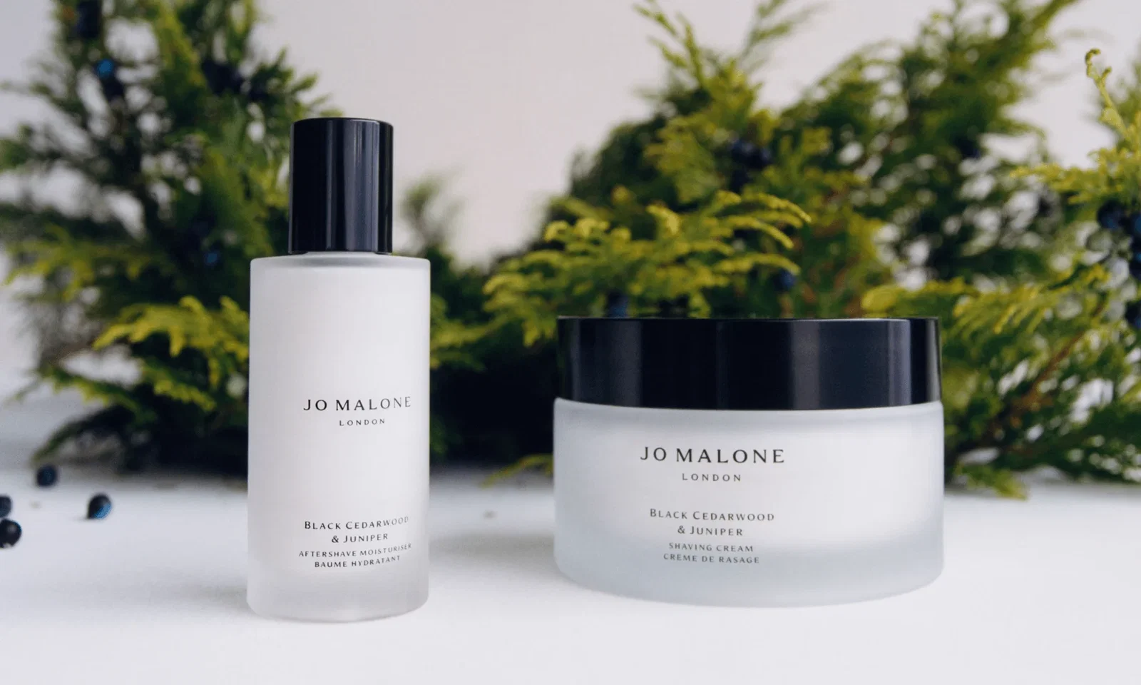 Jo Malone aftershave moisturiser and shaving cream on a smooth white surface, with blurred greenery in the background.