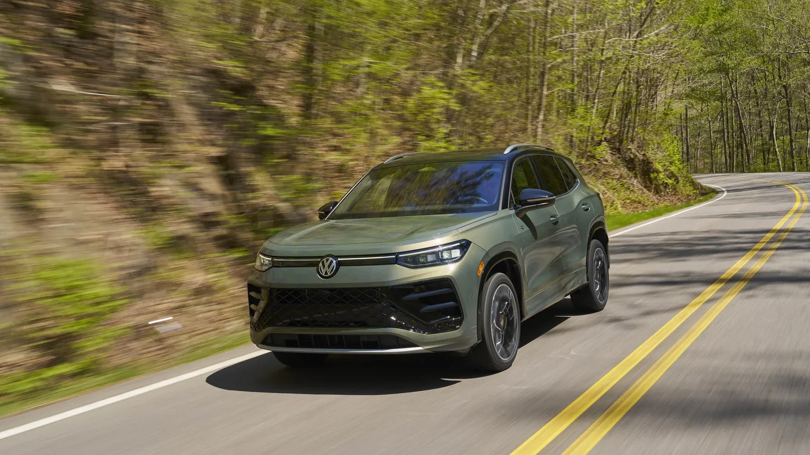Green Volkswagen Tiguan SEL R-Line SUV speeds along a winding forest road, motion blur in the trees, with a double-yellow center line and curved pavement under bright daylight.