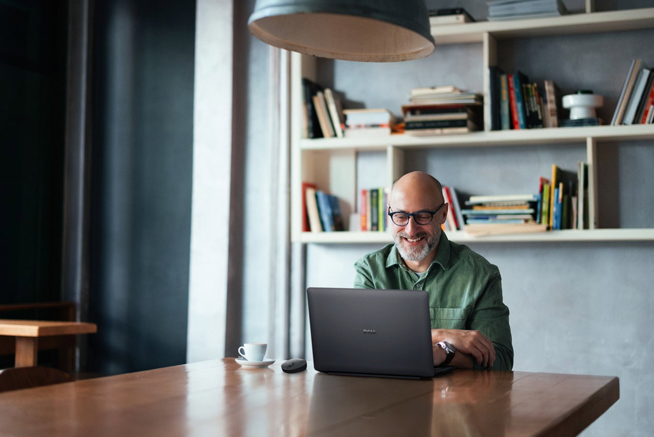 A man sits at a wooden desk in a charming workspace, working on his laptop and enjoying coffee.