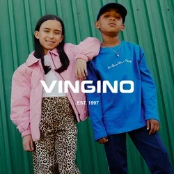 Twee kinderen staan op gras voor een groene metalen muur; de ene draagt een roze jas en luipaardbroek, de andere een blauwe trui en donkere broek.