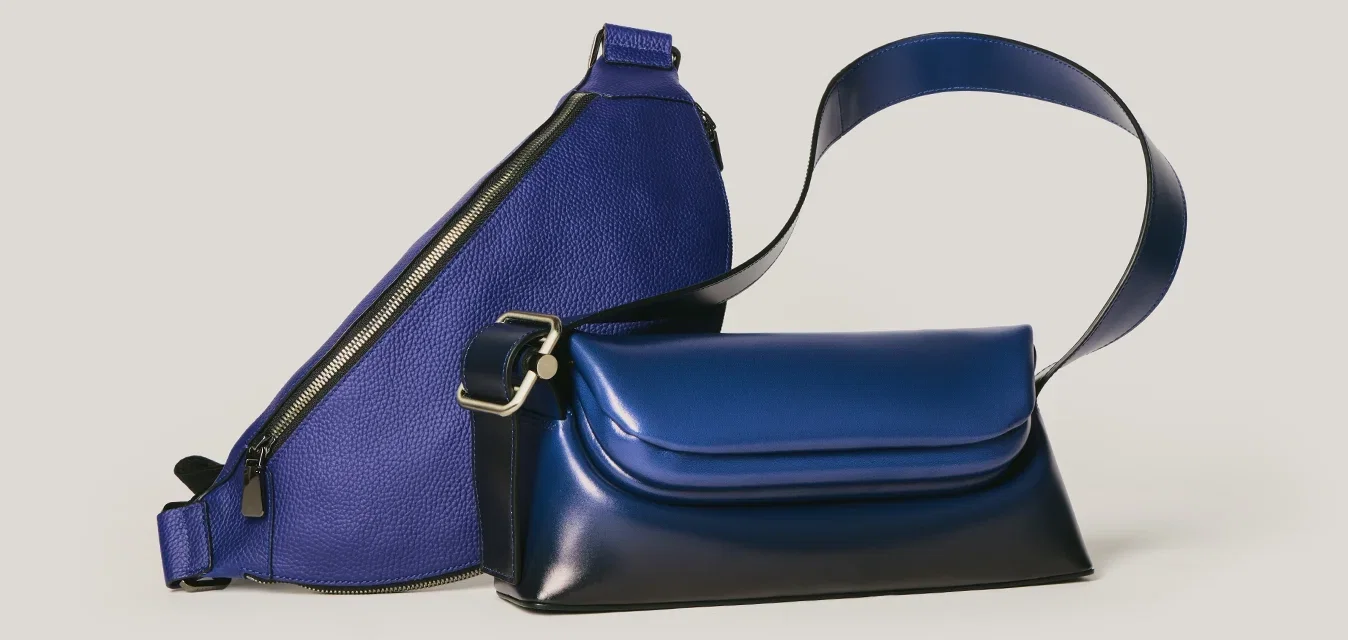 Twee blauwe tassen: één driehoekige crossbodytas met ritssluiting, één rechthoekige handtas met voorklep en brede riem op een lichte achtergrond.