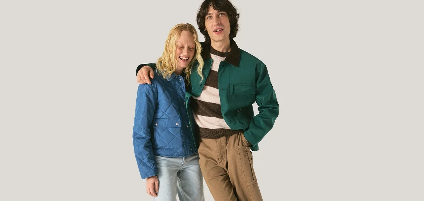 Zwei junge Erwachsene stehen nahe beieinander; einer trägt eine blaue Jacke und helle Jeans, der andere eine grüne Jacke und ein gestreiftes Hemd mit braunen Hosen.
