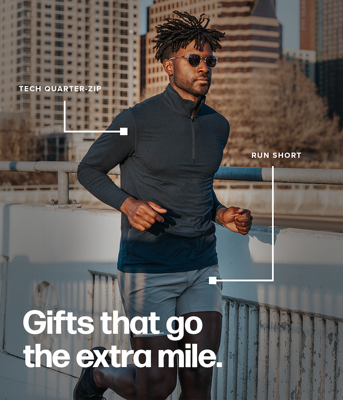 A man jogs outdoors on a concrete bridge with a city skyline behind him. White labels point to his navy blue tech quarter-zip and grey run shorts. Text reads: 'Tech quarter-zip. Run short. Gifts that go the extra mile.'