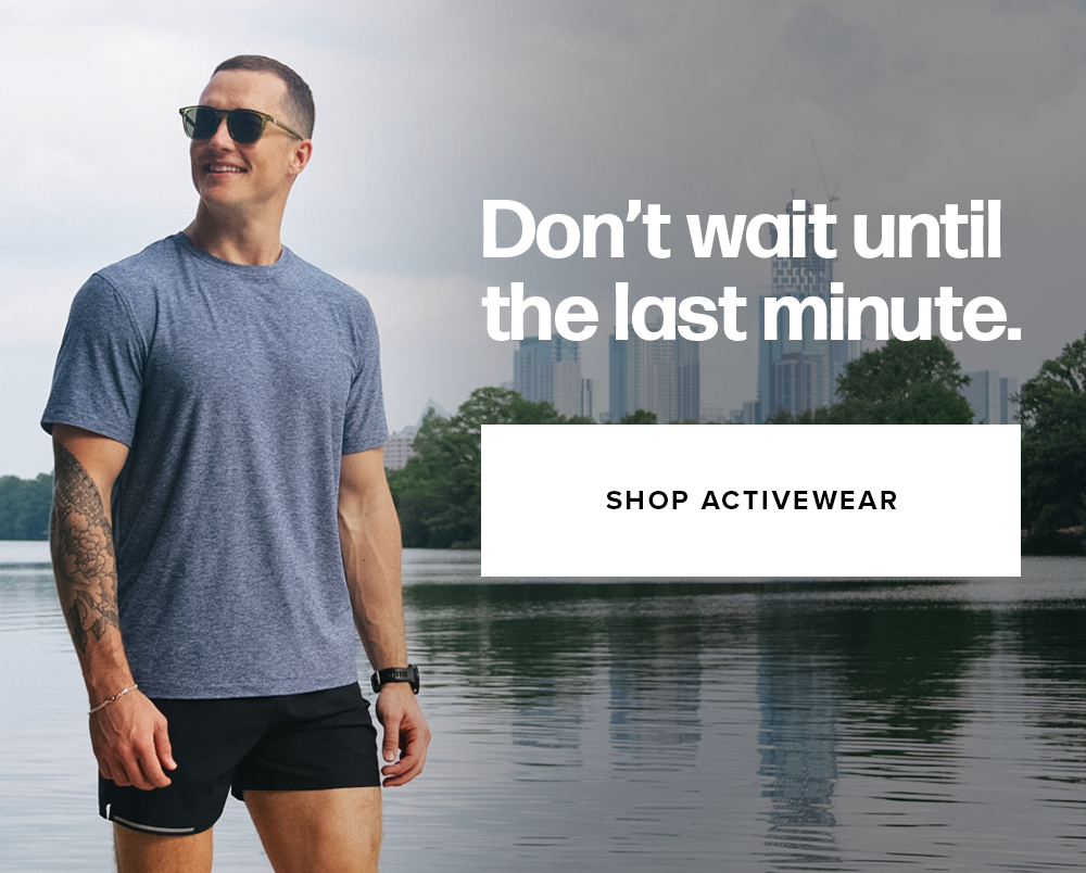 A man wearing sunglasses, a heather blue t-shirt, and black athletic shorts stands near a riverbank with a city skyline in the background. Text reads: 'Don’t wait until the last minute. Shop activewear'
