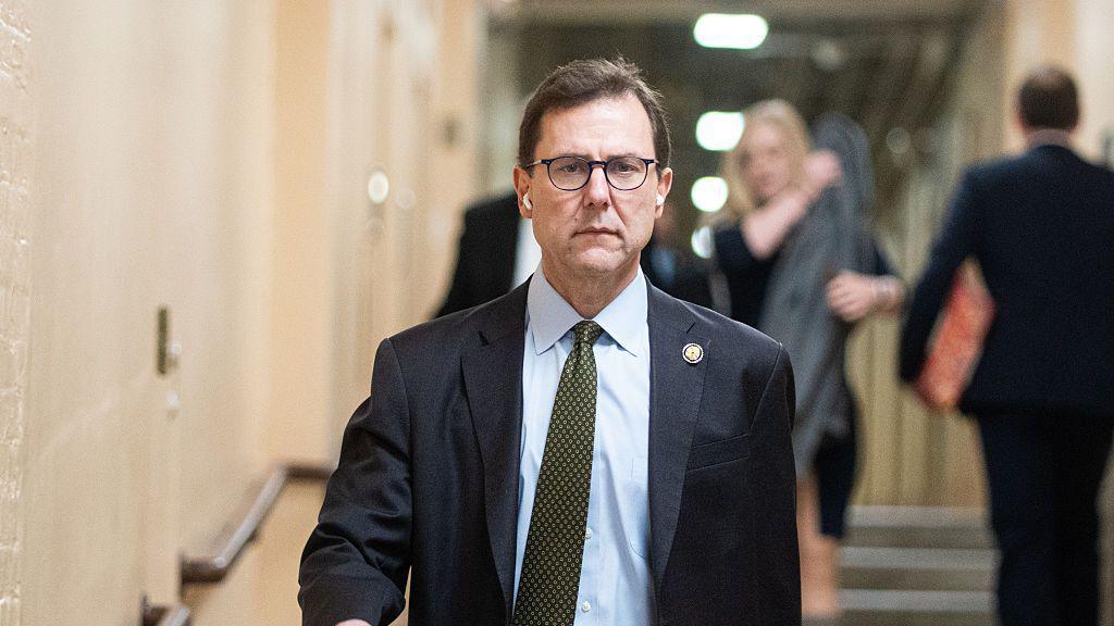 Republican Congressman Tom Kean Jr is pictured candidly walking down stairs wearing a suit and green tie, with white earphones in. The people in the background are slightly blurred.