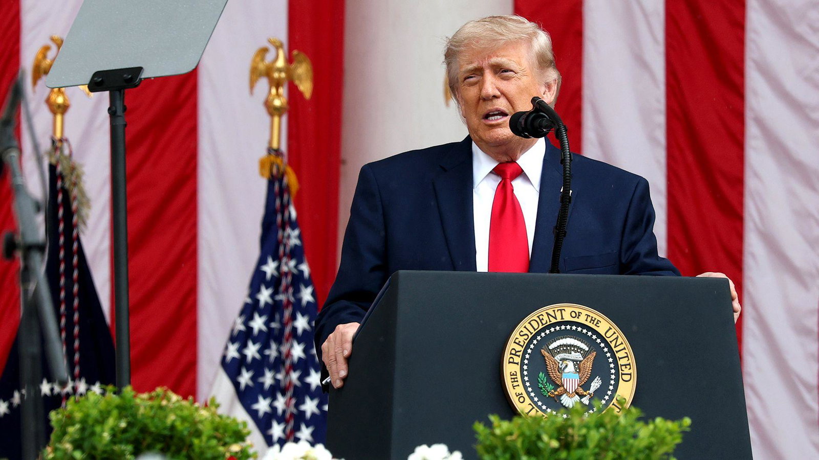 Donald Trump stands behind a lectern with a plaque reading 'President of the United States' with the US eagle seal, as he speaks into a microphone while wearing a dark suit and red tie against the backdrop of the US flag.