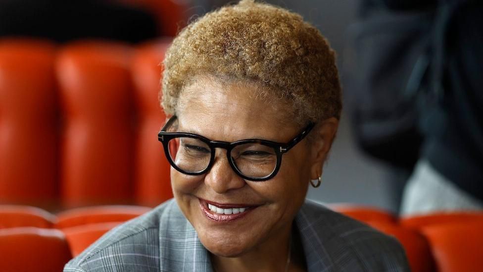 Karen Bass smiles broadly and wears a grey, checked suit. The background, slightly out of focus, appears to be red seating.