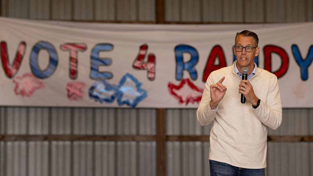 Randy Feenstra speaks into a microphone during a campaign event in Iowa. He wears glasses, jeans and a beige long-sleeved top. A banner that reads ‘Vote 4 Randy’ in blue and red letters is in the background.