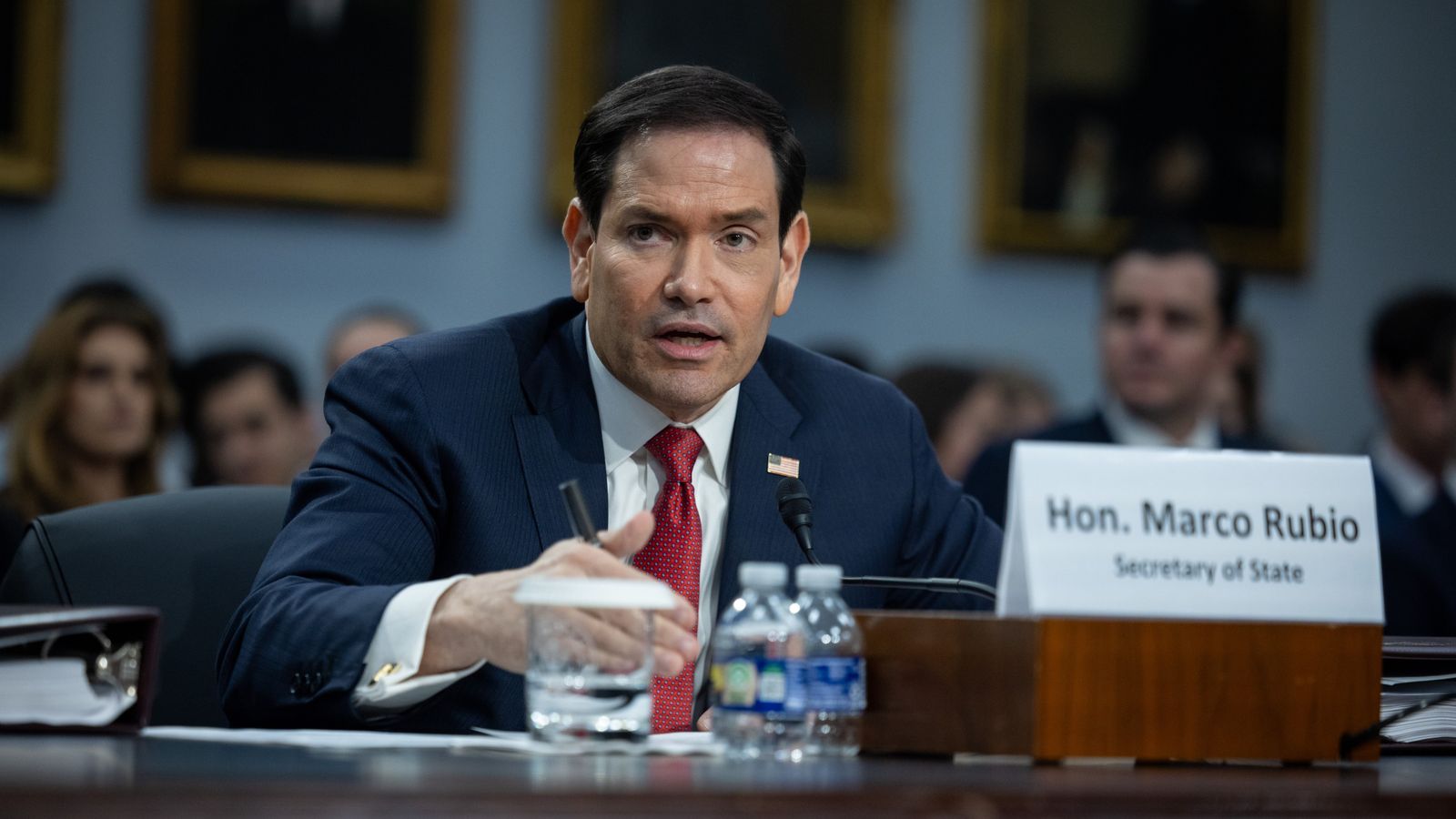 Marco Rubio sits behind a desk and speaks. There is a crowd sat behind him, slightly blurred. There are small bottles of water ont he desk and a paper sign with his name.