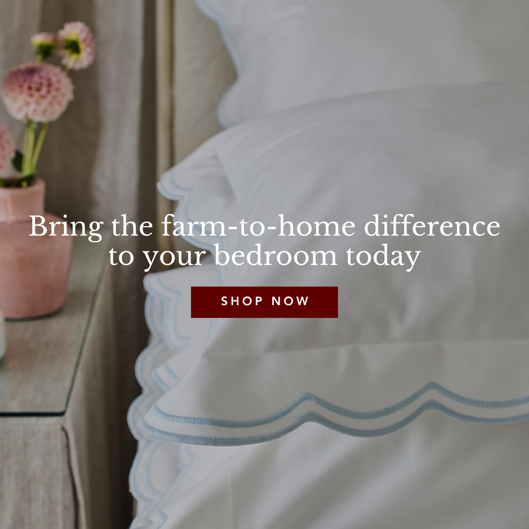 A neatly made bed with white bedding featuring light blue scalloped edges. A vase of pink dahlias sits on a glass bedside table next to the bed.