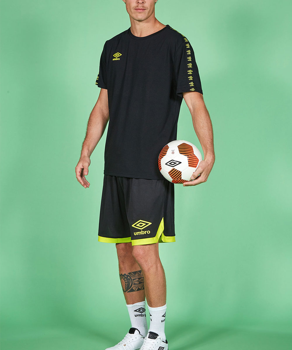 Man holding football wearing black Umbro training kit with neon trim, white socks and calf tattoo on green backdrop