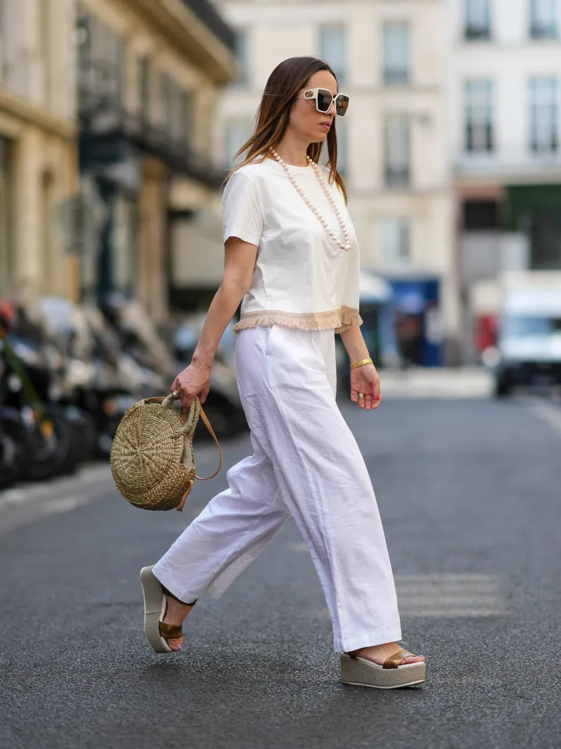 how to style espadrilles - PARIS, FRANCE - MAY 10: Maria Rosaria Rizzo wears white sunglasses, a white pearls long necklace, a white latte t-shirt with fringed borders, a beige wickers handbag from Guerlain, a gold large bracelet, gold rings, high waist white wide legs linen pants, gold shiny leather strappy with wicker wedge heels sandals, during a street style fashion photo session, on May 10, 2022 in Paris, France. (Photo by Edward Berthelot/Getty Images)