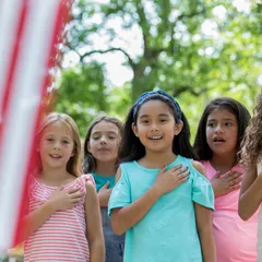 Diverse group of school friends are holding hands over hearts and reciting the Pledge of Allegience to the American flag. Asian American elementary age girl is standing at front of group.