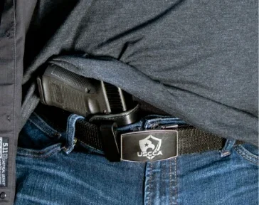 Close-up of a pistol within an AIWB holster, tucked inside someone's blue jeans. The shirt is pulled up slightly to show the grips of the gun and the carrier is wearing a USCCA belt buckle.