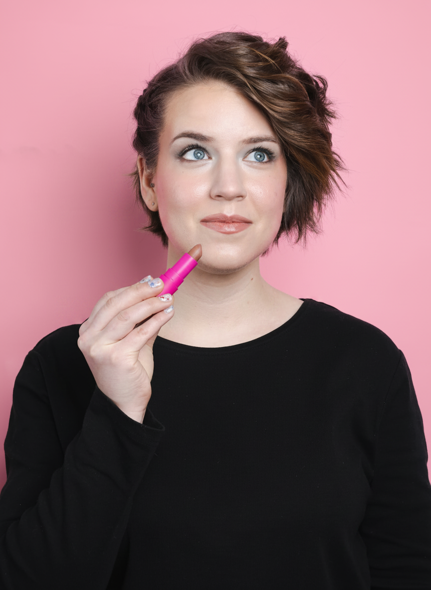 A woman with blue eyes and short brown hair applies lipstick with a pink tube.