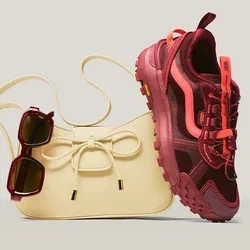 Beige Handtasche mit Schleifendetail, dunkle Sonnenbrille, die am Riemen hängt, und ein roter und pinkfarbener sportlicher Sneaker, der an der Tasche lehnt.