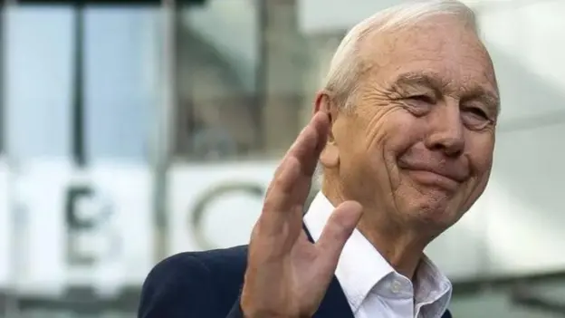John Humphrys waves goodbye outside BBC's New Broadcasting House. He has white hair and is wearing a white shirt with a navy blazer.