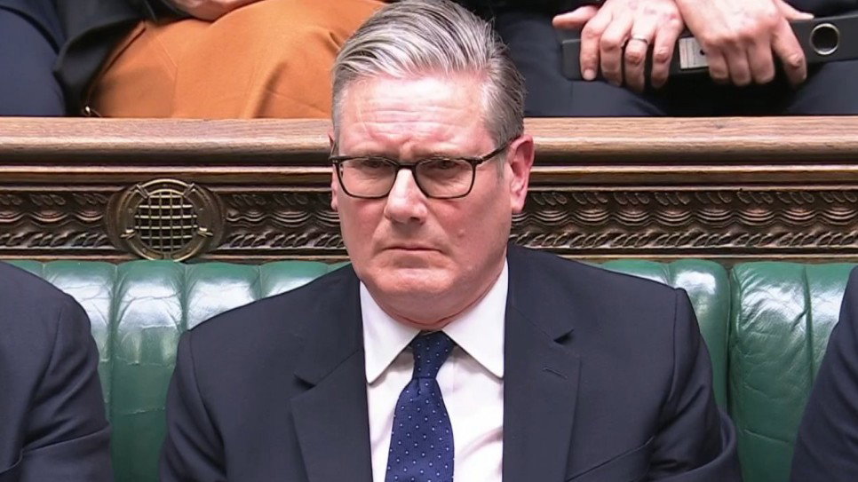 Prime Minister Sir Keir Starmer reacts to Conservative Party leader Kemi Badenoch at PMQs. He is sitting on a green bench in the House of Commons, wearing a black suit, a white shirt and a blue tie. His brow is furrowed.