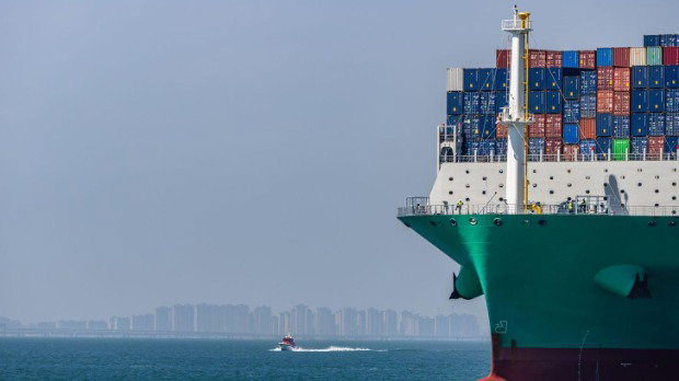 A green shipping container vessel appears on the right of the image, in front of a large cityscape in the background.