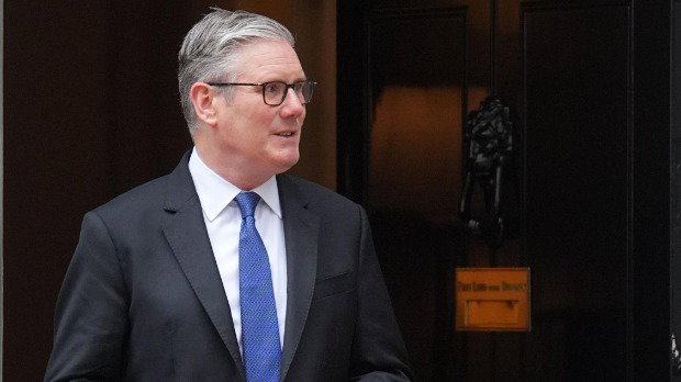 Keir Starmer stands at the door of Downing Streetm wearing a dark suit, blue tie and glasses.