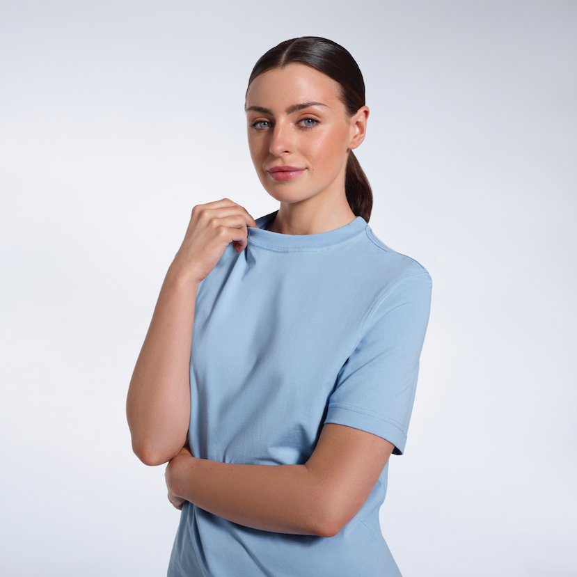 A woman looking into the camera touching her collar, modelling a light blue organic cotton t-shirt.