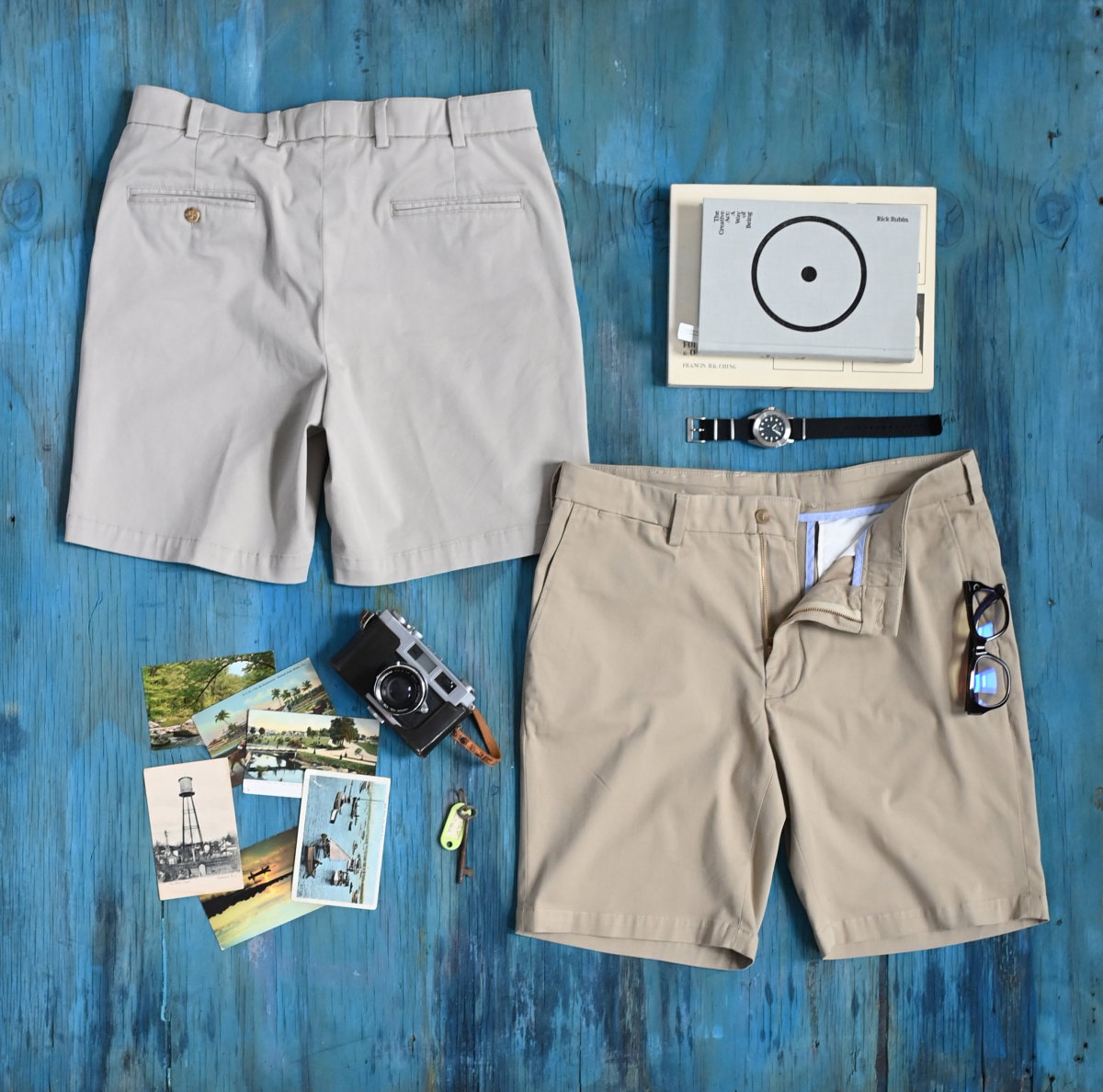 Two pairs of Bills Khakis Travel Twill shorts lay on ocean blue painted wood surface alongside vintage on the go items like books, keys, photos, watch and books