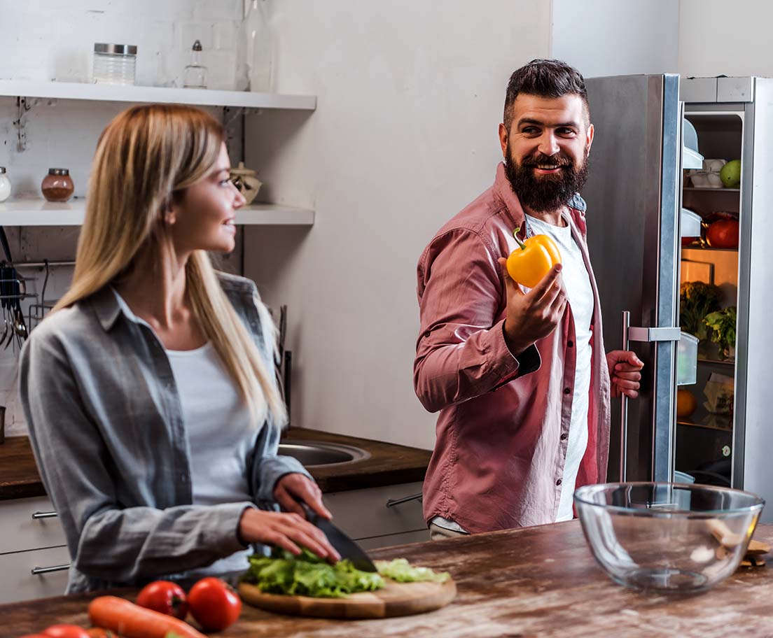 A man holds a yellow bell pepper from an open refrigerator while a woman chops fresh vegetables in the kitchen