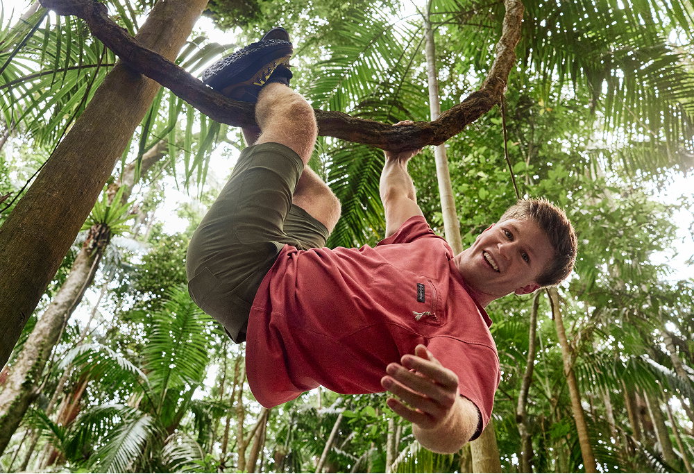 A man climbing a tree, wearing a red short sleeve shirt, green shorts, and brown hiking boots.