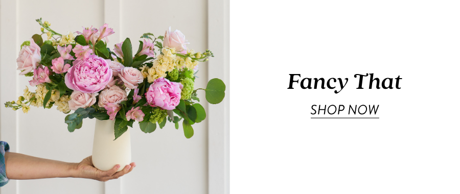 A hand presents a vase arrangement with pink peonies, pale pink flowers, green hydrangea, and greenery next to text reading “Fancy That” and “Shop Now.”