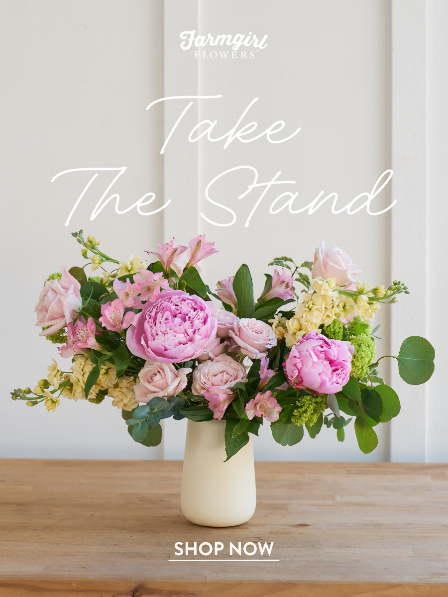 Farmgirl Flowers vase arrangement with pink peonies, pale pink and yellow flowers, green hydrangea, and greenery displayed on a wooden table, with white overlay text reading “Take The Stand” and “Shop Now.”