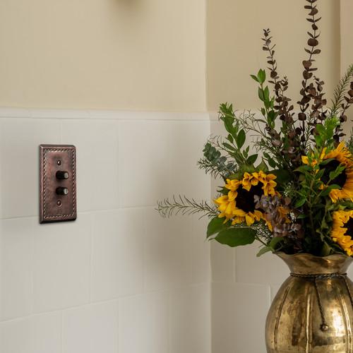 A hammered copper push button plate on a white tile wall next to a vase.