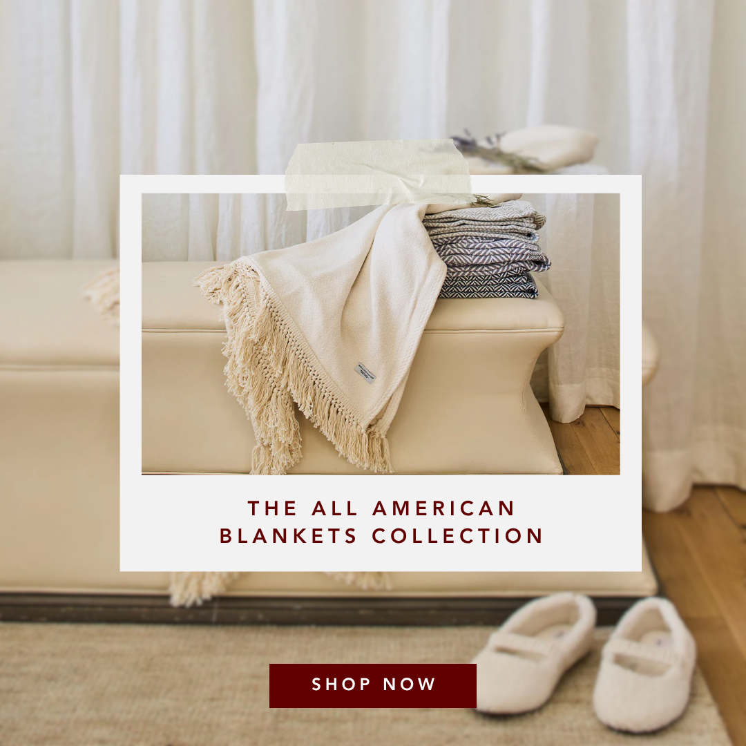 Cream fringed blanket and stacked patterned throws on a beige ottoman, with white slippers on a rug in a cozy room.