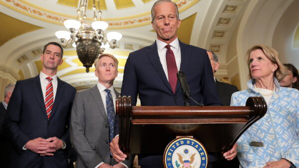Senate Majority Leader John Thune (R-SD) stands at a podium and speaks during a news conference. Several people stand behind him. 