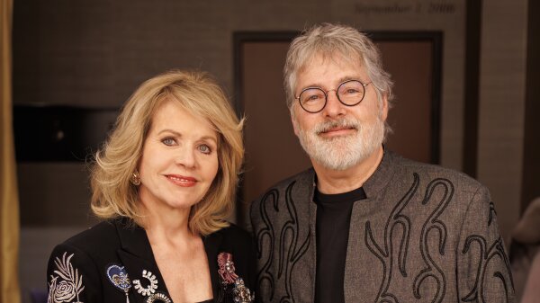 Soprano Renée Fleming and banjo player Béla Fleck from the shoulders up. Fleming has blonde hair and is wearing a black top. Fleck has round-shaped glasses and a white beard and hair. 
