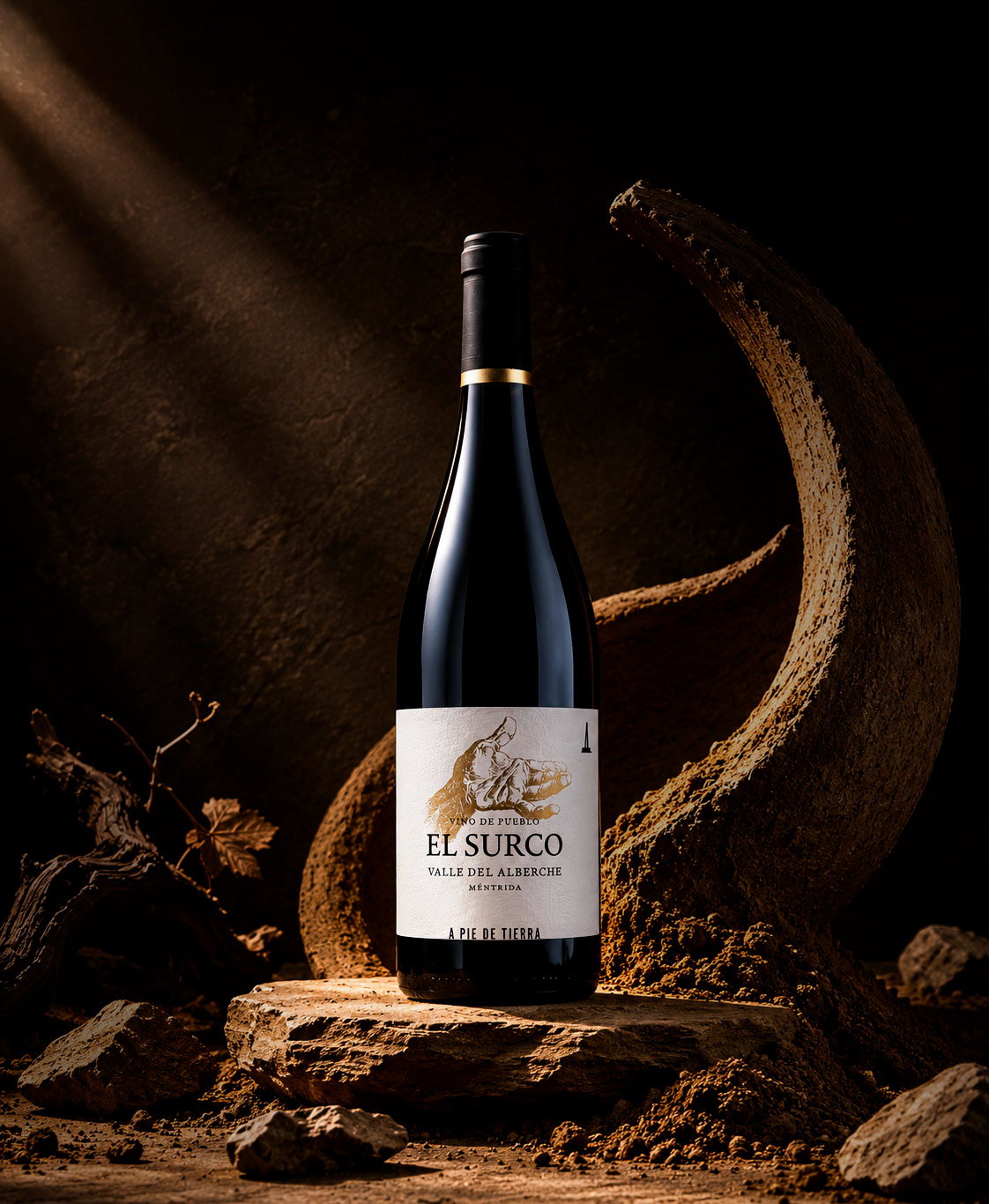 Bottle of El Surco red wine on a rustic stone pedestal amid dry vines and a curved earth formation, lit by a warm spotlight