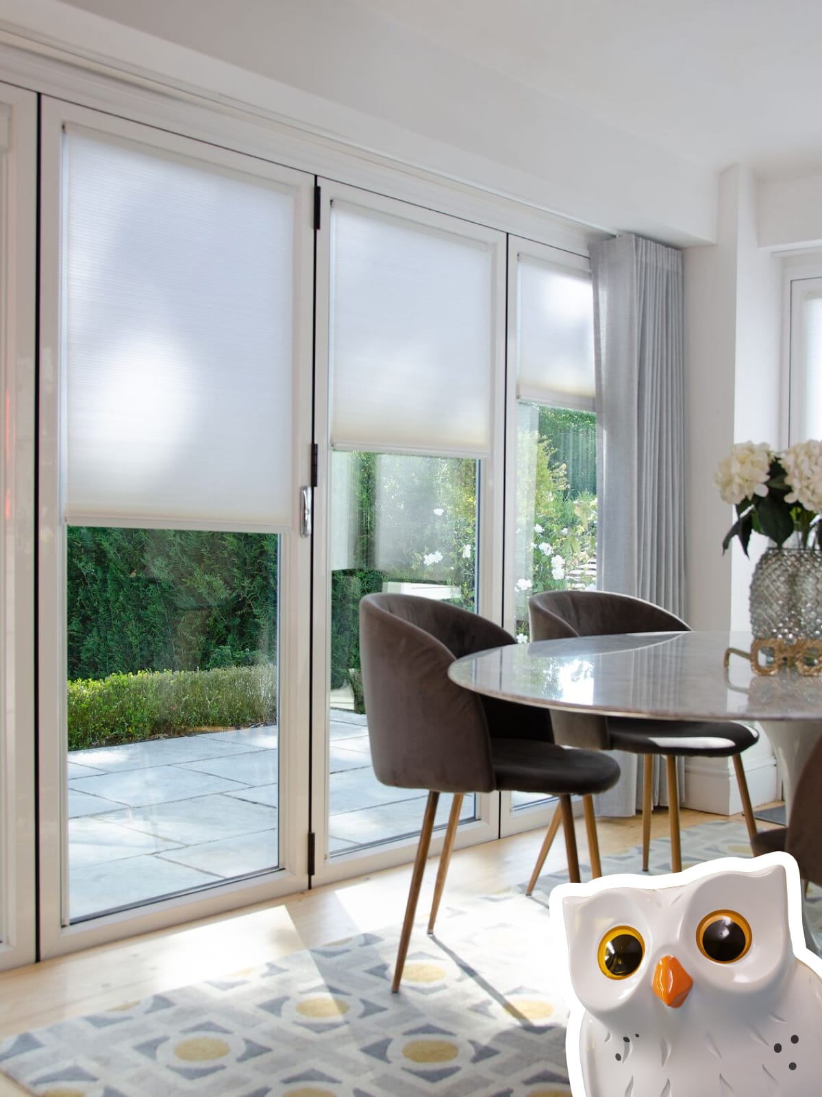 INTU Micro Hive blinds in kitchen diner bifold doors with grey voile curtain pushed over to one side. Table and chairs positioned in front with a vase in the centre. 