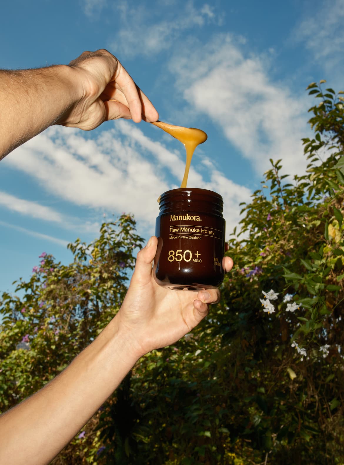A hand holding a dark amber jar of Manukora Raw Mānuka Honey 850+ MGO while another hand drizzles honey from the jar against a bright blue sky and lush green foliage background.
