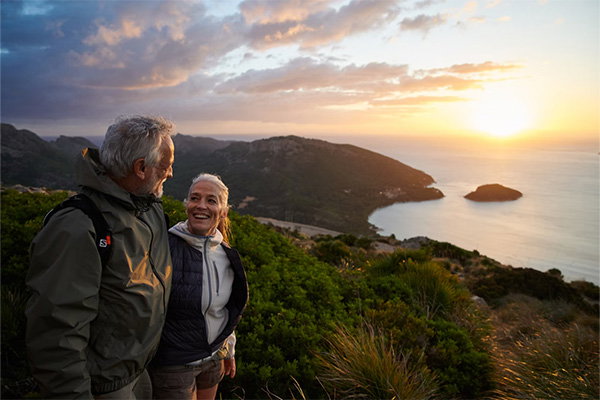 Zwei Wandernde stehen auf einem Hügel und blicken bei Sonnenuntergang auf eine Küstenlandschaft mit Meer.