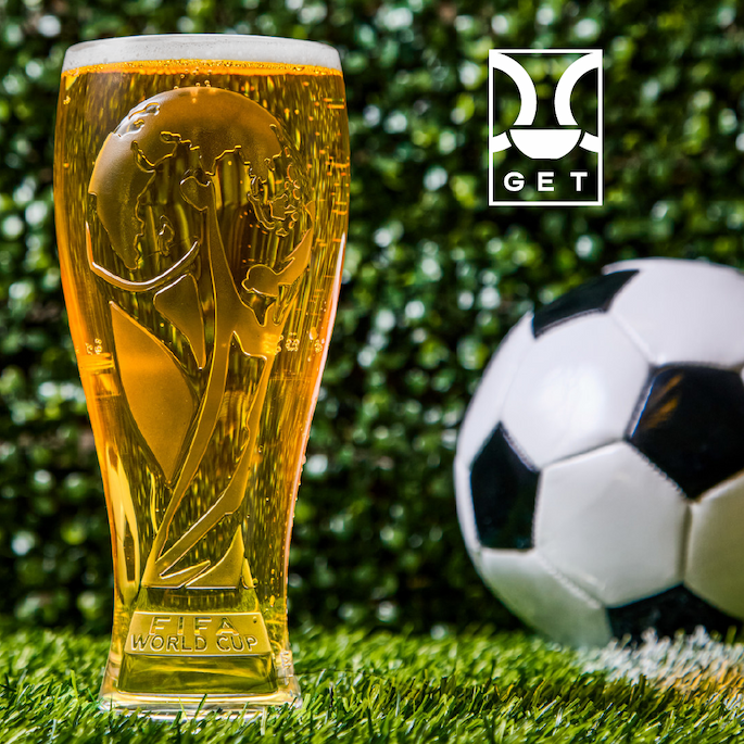 GET branded beer glass shaped like a soccer trophy filled with golden beer, set on artificial turf beside a black and white soccer ball.