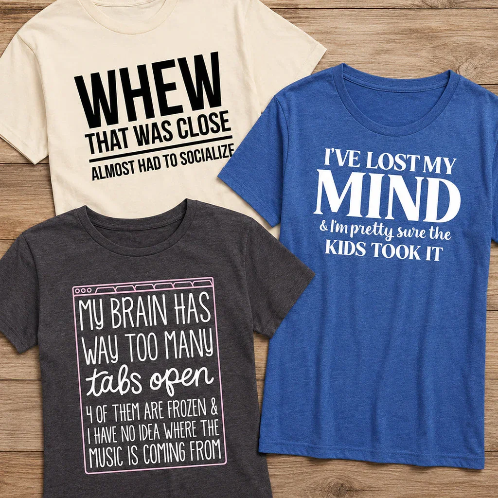 Three humorous graphic t-shirts displayed on wood background featuring funny parenting and life quotes in various colors.