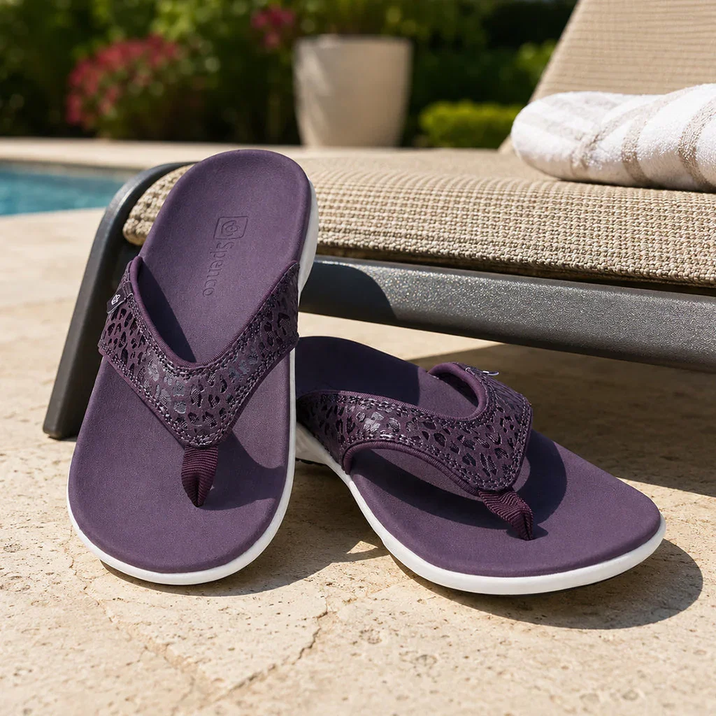 Pair of purple Spenco flip-flops displayed poolside on concrete deck with lounge chairs and garden in background.