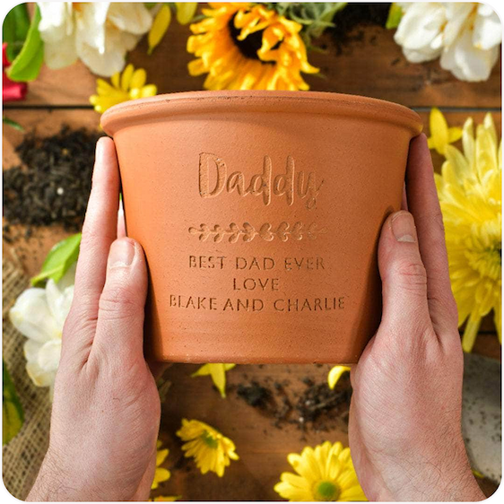 A pair of hands holding an engraved terracotta herb pot with yellow flowers in the background