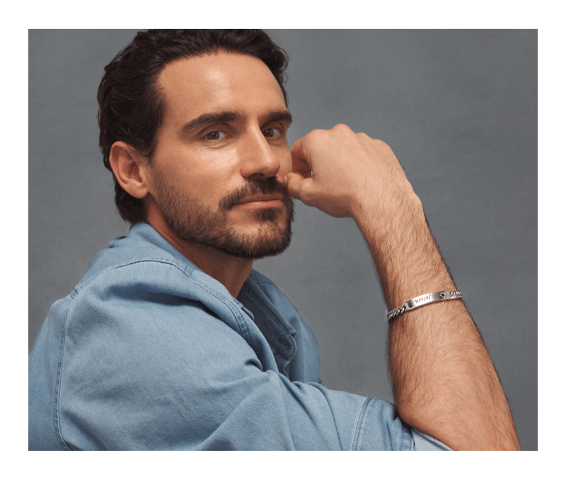 Male model in blue shirt wearing a silver curb chain ID bracelet engraved with Julian and green birthstone star accent.