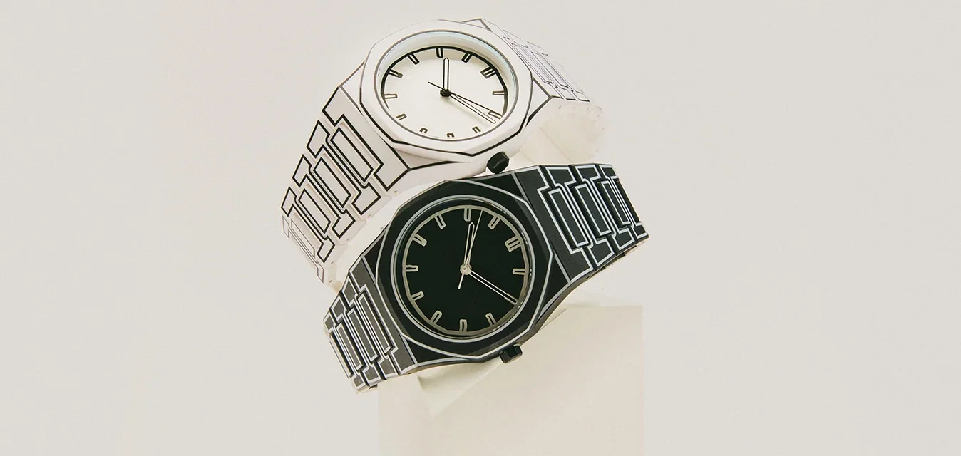 Ein Paar Armbanduhren, eine schwarze und eine weiße, mit geometrisch gemusterten Armbändern, auf einem weißen Ständer vor einem hellen Hintergrund präsentiert.