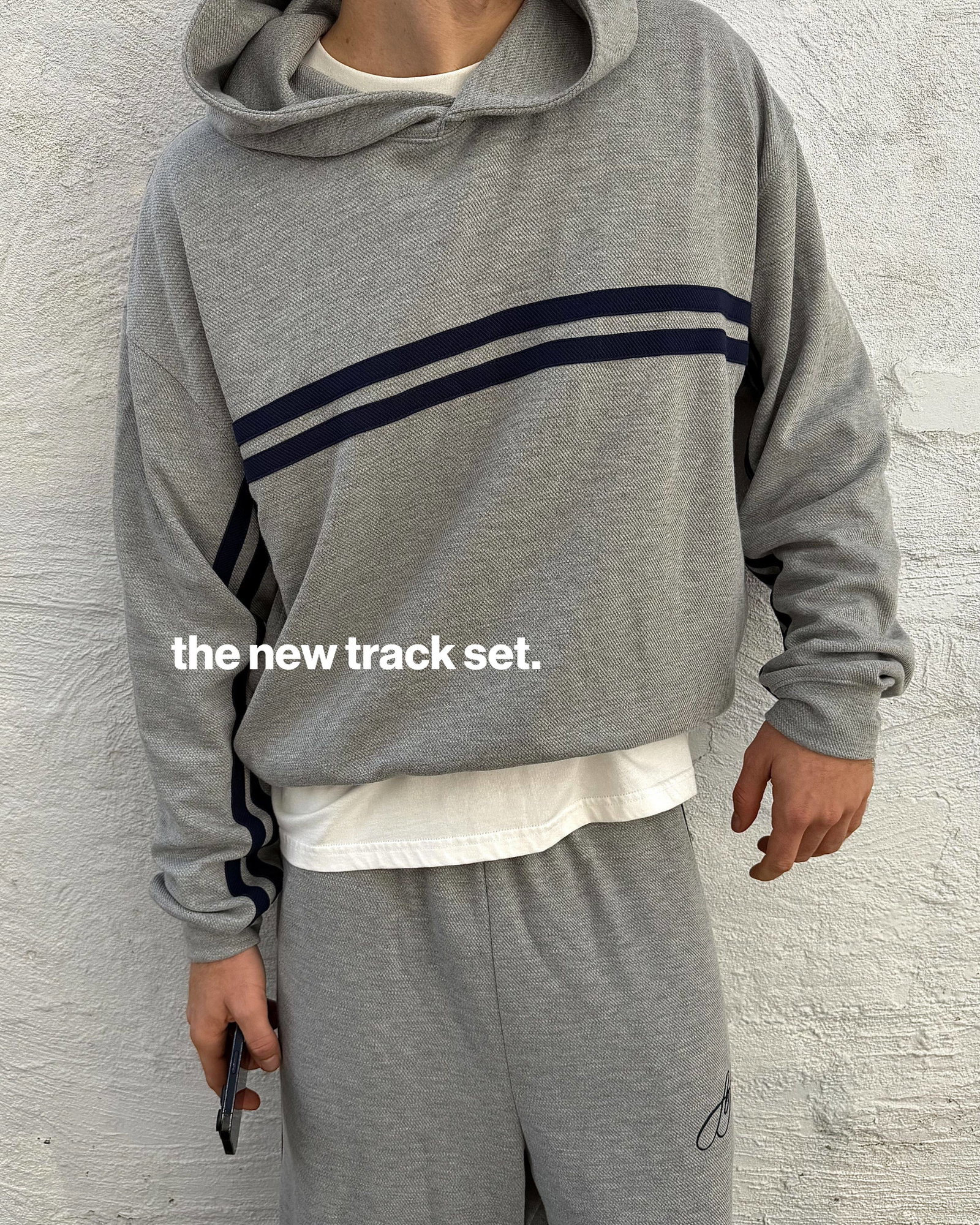 A medium shot shows a person wearing a grey hooded track set with navy blue stripes on the sleeves and chest, layered over a white t-shirt, with the t...