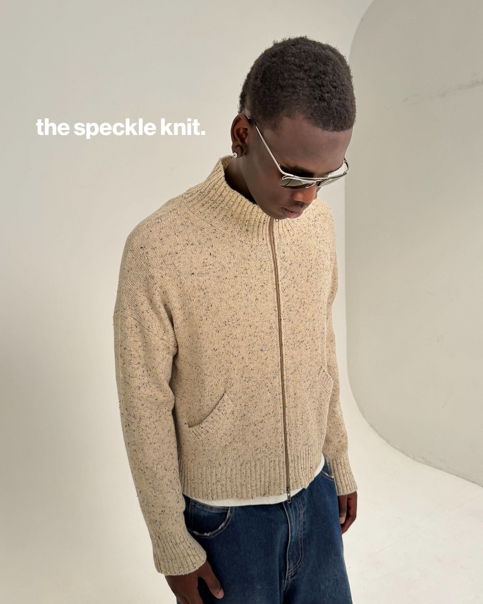 A medium shot captures a person wearing a beige speckled knit zip-up sweater over a white t-shirt and dark wash jeans, with silver-rimmed sunglasses o...