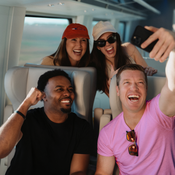 A group of four friends pose for a selfie while traveling on a modern train. Two people sit in the foreground seats, with one holding up a smartphone to capture the moment. Two more friends lean in from the row behind, one wearing a red cap. Large windows reveal a blurred landscape outside, emphasizing motion and travel. The scene conveys a casual, social atmosphere during a shared trip.