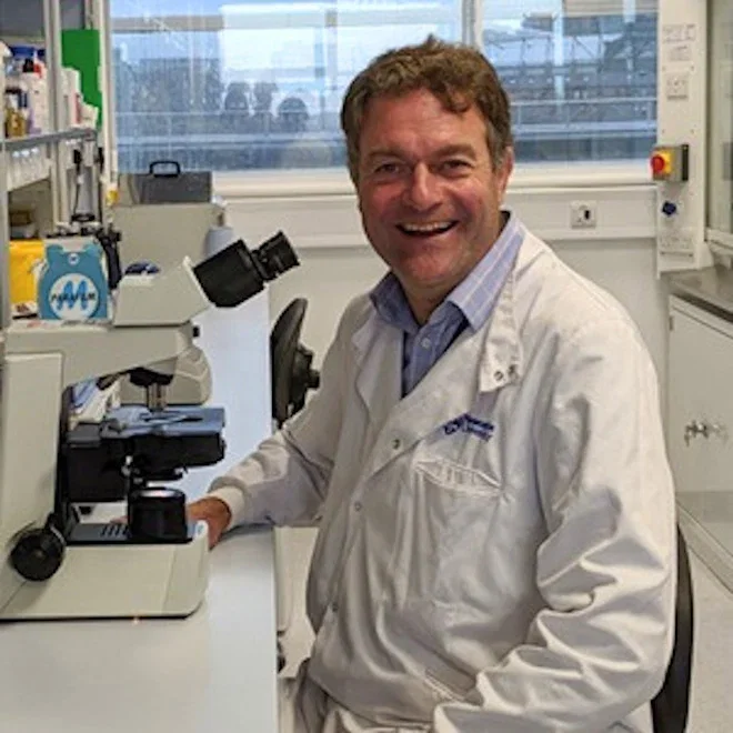 Image of Dr Frederick van Delft, an Action-funded researcher. He is sat in a lab next to a microscope. He is wearing a white lab coat and smiling.