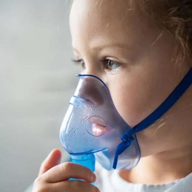 Image is a side-on, close-up photo of a young girl, with a nebuliser over her nose and mouth..